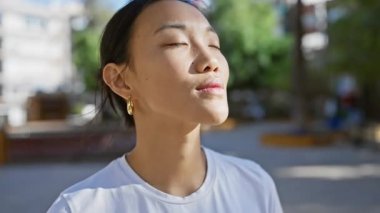 Genç Asyalı bir kadın gözlerini kapar ve sakin bir şehir parkında güneşin tadını çıkarır..