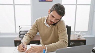 Genç İspanyol bir adam birden fazla iş yapıyor. Ofis masasında notlar yazarken telefonda konuşuyor. Modern bir çalışma alanında profesyonelliği somutlaştırıyor..