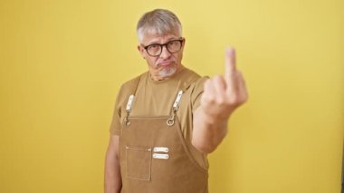 Impudent middle-age grey-haired man in glasses and apron standing, showcasing a rude 'fuck you' hand gesture. angry expression over isolated yellow wall background
