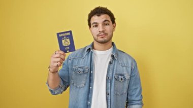 Şaşırmış genç bir Arap adam, şok ve güvensizlik içinde ağzı bağlı, Emirlik pasaportunu tutuyor, sarı arka planda şaşkınlıktan donmuş.