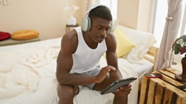 Parlak bir odada yatakta otururken kulaklık takan yakışıklı Afro-Amerikalı adam.