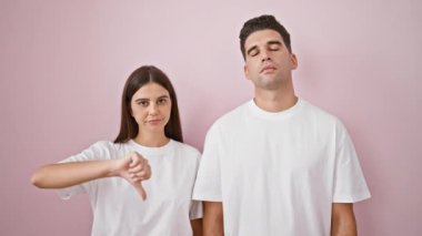Hoşnutsuz bir kadın, pembe bir duvara dayanmış bir adamın yanında, memnuniyetsizliğini gösteren bir başparmağını kaldırır..