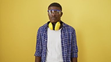 Güvenilir Afro-Amerikan adam, ciddi bir ifadeyle, güvenlik gözlüğü takıyor. Mükemmel bir şekilde dengeli duruyor, doğal ve basit bir görünümü somutlaştırıyor. Sarı arkaplana karşı izole bir figür.