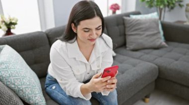 Modern oturma odasında akıllı telefon kullanan İspanyol genç kadın..