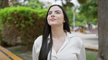 Esmer, sıradan gömlekli İspanyol bir kadın yeşil bir parkta poz veriyor. Güzellik ve sükunet hissi veriyor..