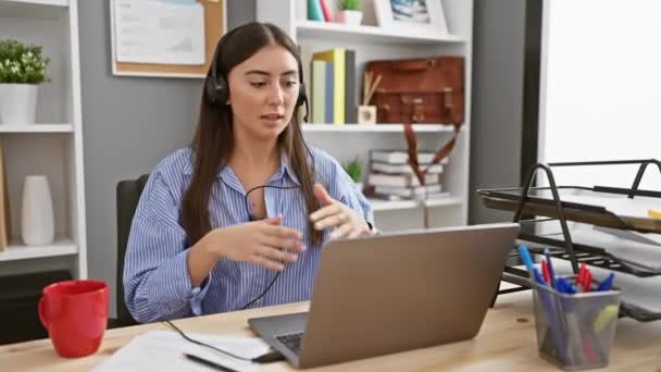 一位带着耳机的年轻的惊慌失措的女人在办公室桌上带着笔记本电脑和红色杯子进行视频通话 — 图库视频影像