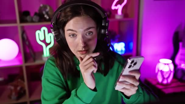 ヘッドフォンを着用している若い女性は 夜間に活気に満ちたネオンライトゲームルームでスマートフォンとやり取りします — ストック動画