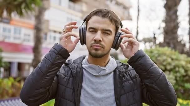 Młody Brodaty Mężczyzna Dostosowuje Słuchawki Stojąc Ulicy Miejskiej Zielenią Tle — Wideo stockowe