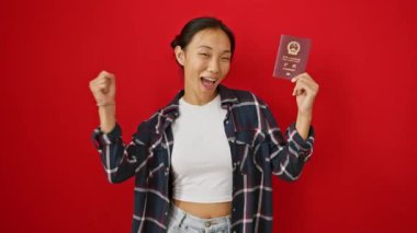 Zafer kazanmış genç Çinli kadın gururla pasaport kaldırıyor, izole edilmiş kırmızı arka plana neşe ve heyecan saçıyor.