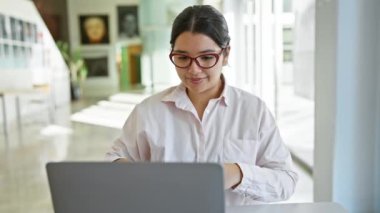 Gözlüklü genç İspanyol kadın modern ofiste dizüstü bilgisayarda çalışıyor. İç mimarisi bulanık..