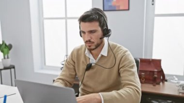 Modern bir ofiste dizüstü bilgisayarda kulaklık takan genç sakallı bir adam yorgun ve bilgisayarını kapatıyor..