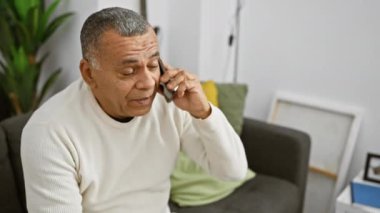 Olgun bir İspanyol adam, rahat ve modern bir oturma odasında akıllı bir telefonla konuşarak, özenli olduğunu ifade eder..