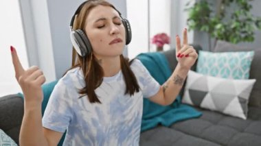 Genç bir kadın, rahat bir oturma odasında kulaklıklarla müzik dinlemekten zevk alır. Elleriyle işaret eder..