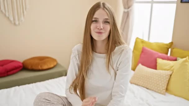 ヤング 魅力的なブロンドの女性 パジャマ 彼女の居心地の良い寝室のベッドに座って フレンドリーな援助と受け入れの兆候として歓迎空の手のジェスチャーを提供 — ストック動画