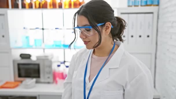 在一个设备齐全的现代化实验室里 一位身穿实验室外套和防护眼镜的笑容满面的年轻女子自信地站在那里 — 图库视频影像