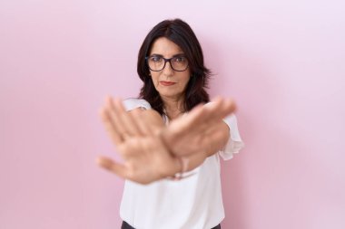 Orta yaşlı İspanyol kadın, günlük beyaz tişört ve gözlük takıyor. Reddedilmiş yüz ifadesi, kollar ve avuçlar üzerinde negatif işaretler, kızgın yüz ifadesi. 