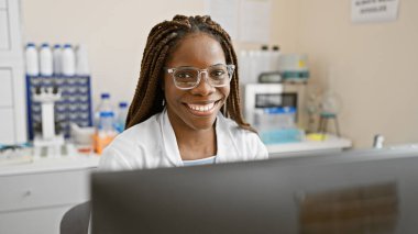 Laboratuvarda laboratuvar önlüğü ve gözlüğü takmış, gülümseyen Afrikalı Amerikalı bir kadının portresi.