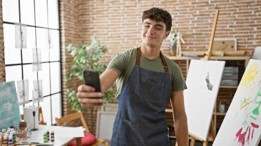 Kendine güvenen genç İspanyol sanatçı, gülümsüyor, resim stüdyosunda unutulmaz bir özçekim yapıyor, akıllı telefondaki resim yolculuğunu kaydediyor.