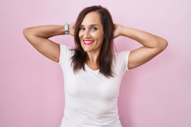 Orta yaşlı esmer kadın pembe arka planda duruyor rahatlıyor ve esniyor, kolları ve elleri başının ve boynunun arkasında mutlu bir şekilde gülümsüyor. 