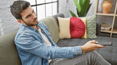 Kot gömlekli İspanyol adam modern bir oturma odasında uzaktan kumanda tutuyor.
