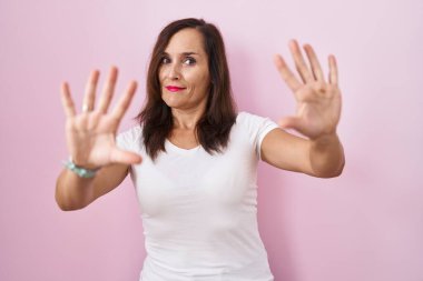 Orta yaşlı esmer kadın pembe arka planda korkmuş ve korkudan dehşete düşmüş halde duruyor. El hareketlerini durdur, şok içinde bağır. panik konsepti. 