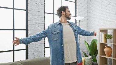 Kot ceket giyen, sakallı, neşeli İspanyol bir adam kollarını geniş bir salonda modern dekorasyonla açıyor..