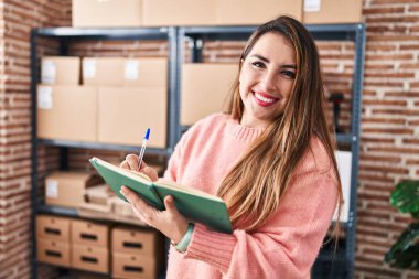 Young beautiful hispanic woman ecommerce business worker writing on notebook standing at office