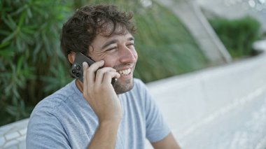 Parkta gülümseyen genç adam telefonda konuşuyor.