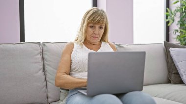 Konforlu evinde, ciddi ve odaklanmış orta yaşlı sarışın bir kadın koltukta oturuyor, tamamen içine kapanık, oturma odasındaki bilgisayarını kullanıyor..