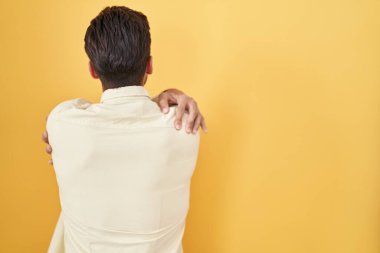 Sarı arka planda dikilen İspanyol genç adam kendine sarılıyor mutlu ve pozitif bir şekilde. Sevgi ve özgüven. 