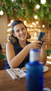 Genç, güzel, İspanyol bir kadın restoranda masada otururken mutlu bir şekilde gülümsüyor.