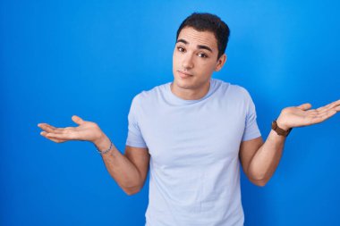 Young hispanic man standing over blue background clueless and confused expression with arms and hands raised. doubt concept. 