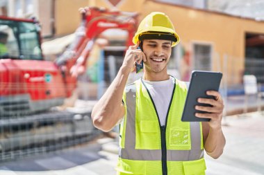 Young hispanic man architect talking on smartphone using touchpad at park