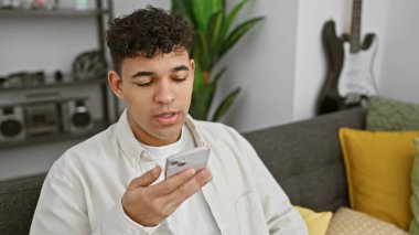 Yakışıklı bir genç adam modern bir oturma odasında rahatça otururken dikkatle akıllı telefonuna odaklanır..