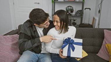 Gülümseyen bir kadın, sıcak bir oturma odasındaki kanepede oturan bir adamdan sevgi ve kutlamaları simgeleyen bir hediye alır..