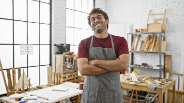 İyi aydınlatılmış bir marangozluk atölyesinde kollarını kavuşturmuş gülümseyen genç bir adam.