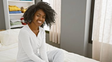 Gülümseyen Afrikalı bir kadın aydınlık yatak odasında rahatça oturuyor, mutluluk ve rahatlama hissi yayıyor..