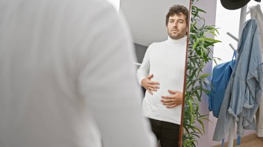 Beyaz kazaklı yakışıklı genç adam modern bir soyunma odasında aynada kendine bakıyor..