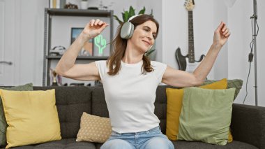Evde kulaklıkla dans eden neşeli bir kadın, rahat bir oturma odasında müziği hissediyor..