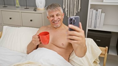 Gri saçlı, elinde kırmızı bardakla gülümseyen adam telefonuyla selfie çekiyor.