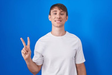 Mavi arka planda duran beyaz tenli sarışın adam iki numaralı parmaklarıyla işaret ederken kendinden emin ve mutlu bir şekilde gülümsüyor.. 