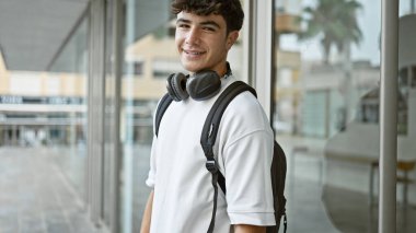Neşeli genç İspanyol erkek öğrenci kampüs hayatının tadını çıkarıyor, kulaklık ve sırt çantası takıyor, üniversitede pozitif enerji ve neşe saçıyor..