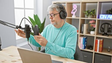 Kulaklık ve mikrofonla donatılmış, deniz mavisi süveteri giymiş olgun bir adam stüdyodan podcast yapıyor..