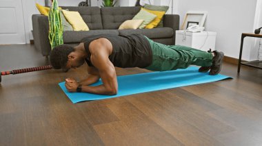 Genç bir Afro-Amerikalı içeride egzersiz yapıyor, temiz bir oturma odasında mavi bir paspas üzerinde tahta yapıyor..