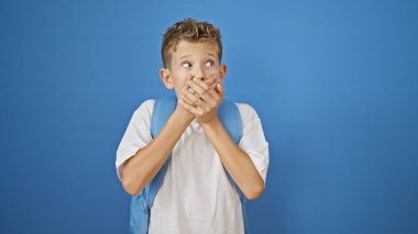 Sevimli sarışın öğrenci şaşkınlık içinde, ağzı kapalı, mavi arka planda çalışma çantasıyla ayakta duruyor.