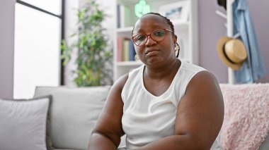 Kanepede oturan Afro-Amerikalı kadın ciddi bir ifadeyle evde oturuyor.