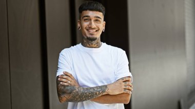 Portrait of a young, confident latin man, sporting a tattoo, cheerfully standing on a city street while expressing joy. his smiling face exudes positivity, his arms crossed in a gesture of confidence. clipart