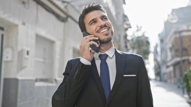 Yakışıklı İspanyol takım elbiseli adam güneşli bir caddede telefonla konuşurken gülümsüyor.