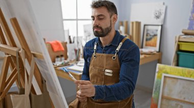 Rahatlamış genç İspanyol bir adam, yakışıklı sakallı bir ressam portresi, resim stüdyosunda fırçalarla resim yapmaya odaklanmış, tuval başyapıtını mükemmelleştiriyor.