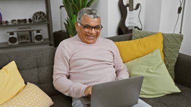 Gri saçlı gülümseyen bir adam, rahat bir şekilde gri bir koltukta renkli yastıklarla oturur ve rahat bir oturma odasında dizüstü bilgisayarda çalışır..
